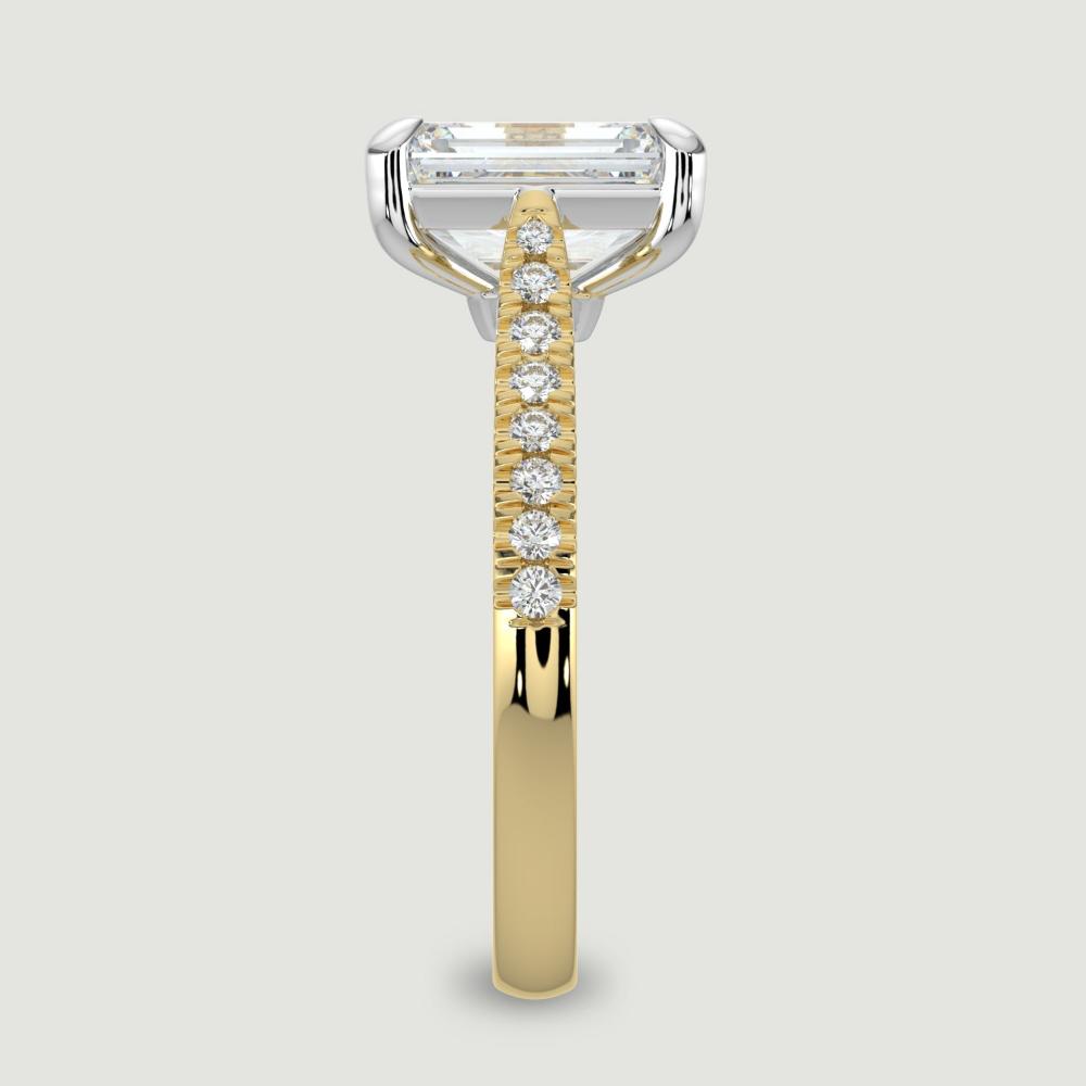 An 18ct yellow gold single stone diamond engagement ring, with micro-pavé set shoulders set with a lab-grown emerald cut diamond held in a platinum four claw setting. Viewed from the edge.