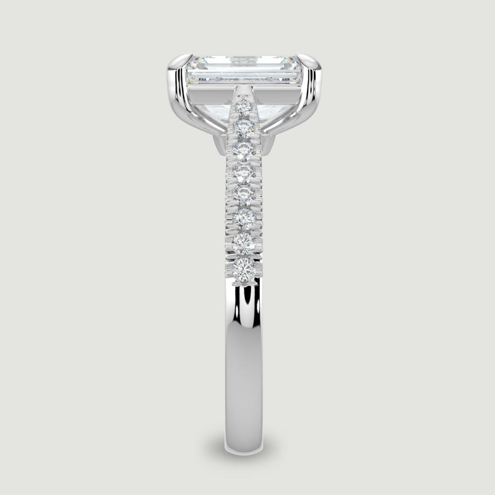 A platinum single stone diamond engagement ring, with micro-pavé set shoulders set with a lab-grown emerald cut diamond held in a four claw setting. Viewed from the edge.