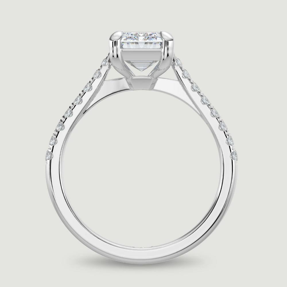 A platinum single stone diamond engagement ring, with micro-pavé set shoulders set with a lab-grown emerald cut diamond held in a four claw setting. Viewed from the side.