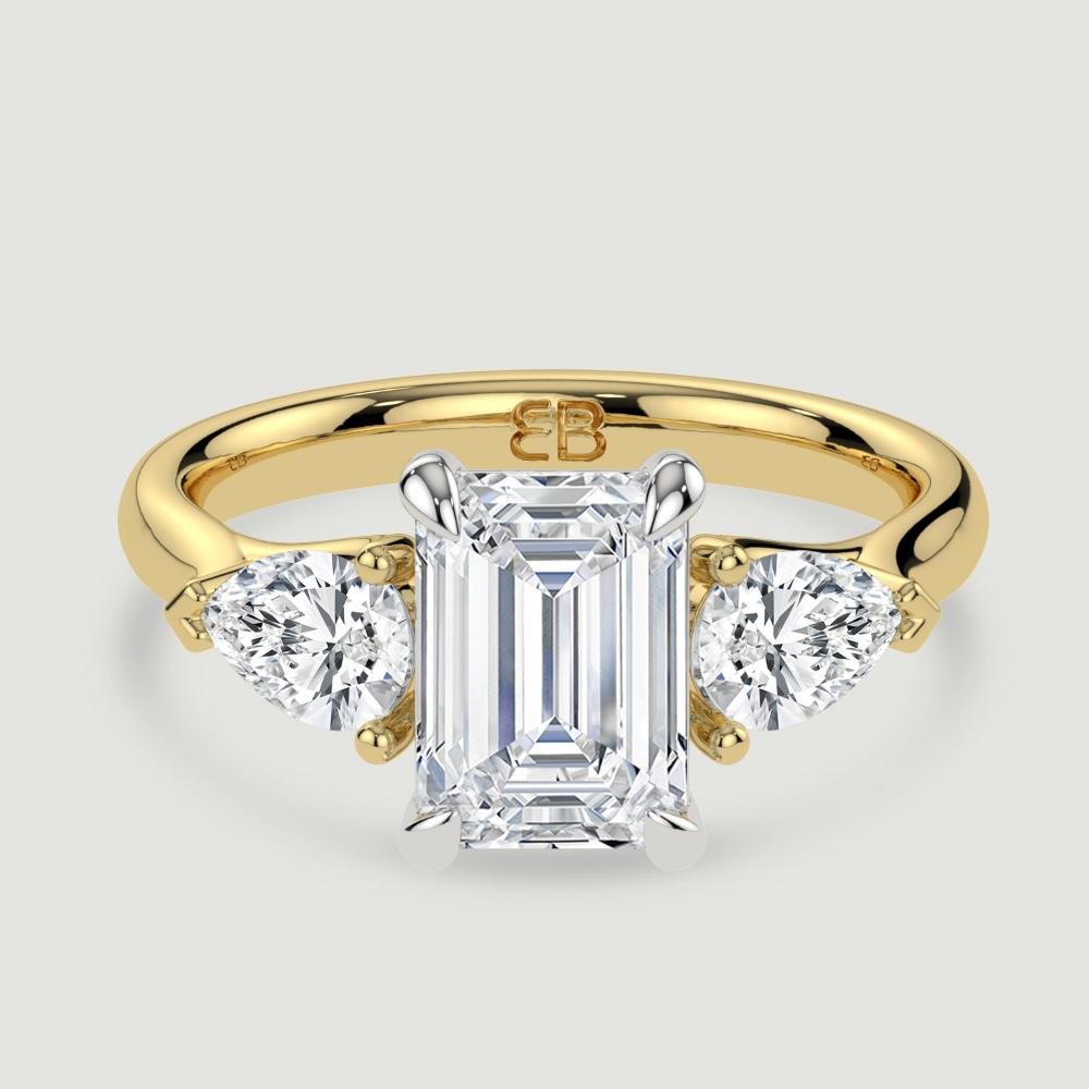 An 18ct yellow gold three stone diamond engagement ring, set with a lab-grown emerald cut diamond centre stone held in a platinum four claw setting flanked by a pear shaped lab-grown diamond on either side, held in three claws. Viewed from above.
