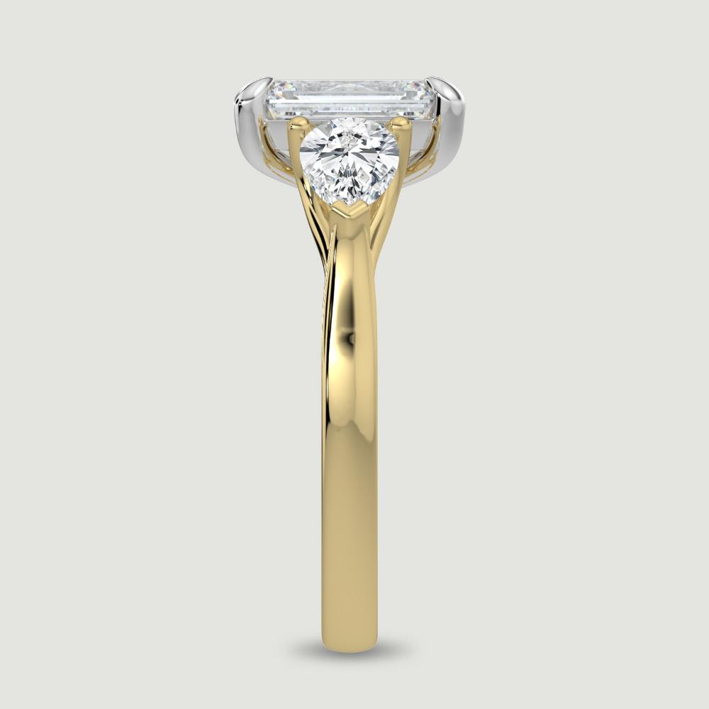 An 18ct yellow gold three stone diamond engagement ring, set with a lab-grown emerald cut diamond centre stone held in a platinum four claw setting flanked by a pear shaped lab-grown diamond on either side, held in three claws. Viewed from the edge.
