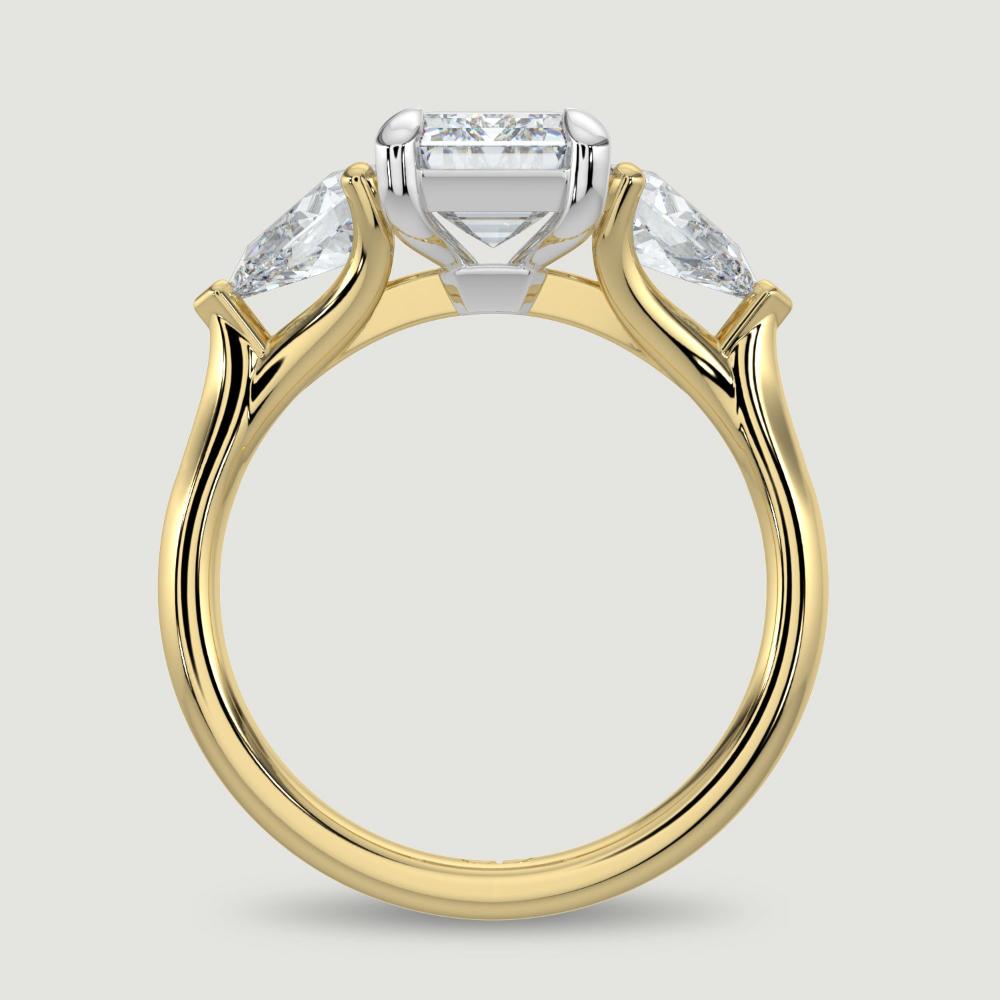 An 18ct yellow gold three stone diamond engagement ring, set with a lab-grown emerald cut diamond centre stone held in a platinum four claw setting flanked by a pear shaped lab-grown diamond on either side, held in three claws. Viewed from the side.