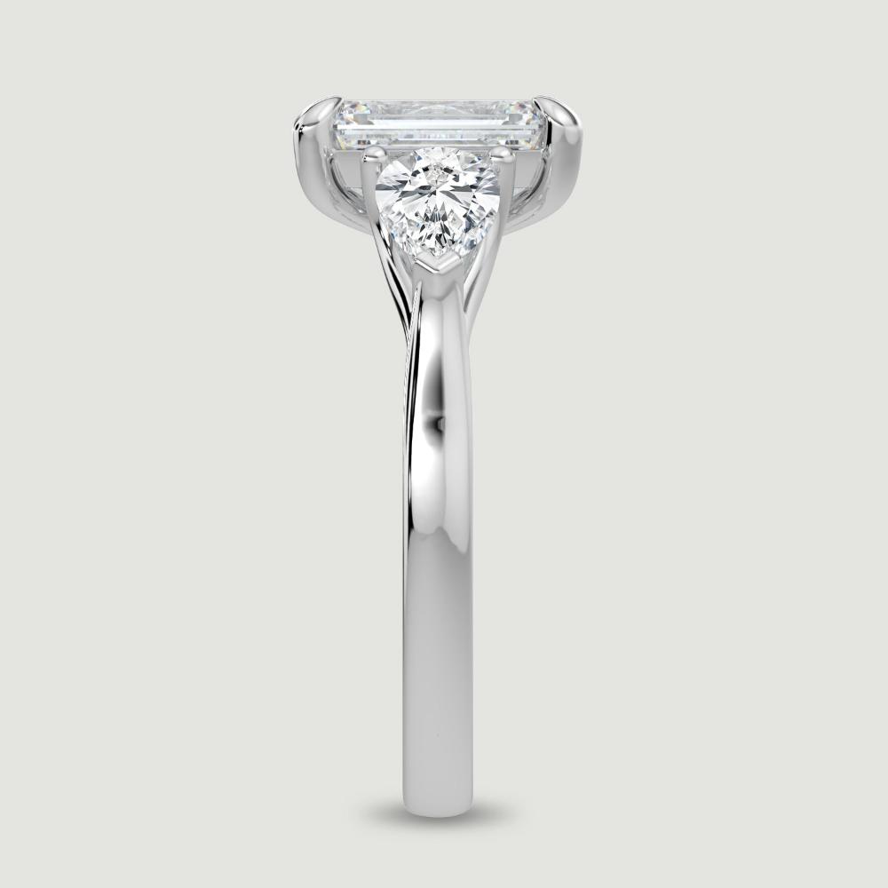 A platinum three stone diamond engagement ring, set with a lab-grown emerald cut diamond centre stone held in a four claw setting flanked by a pear shaped lab-grown diamond on either side, held in three claws. Viewed from the edge.