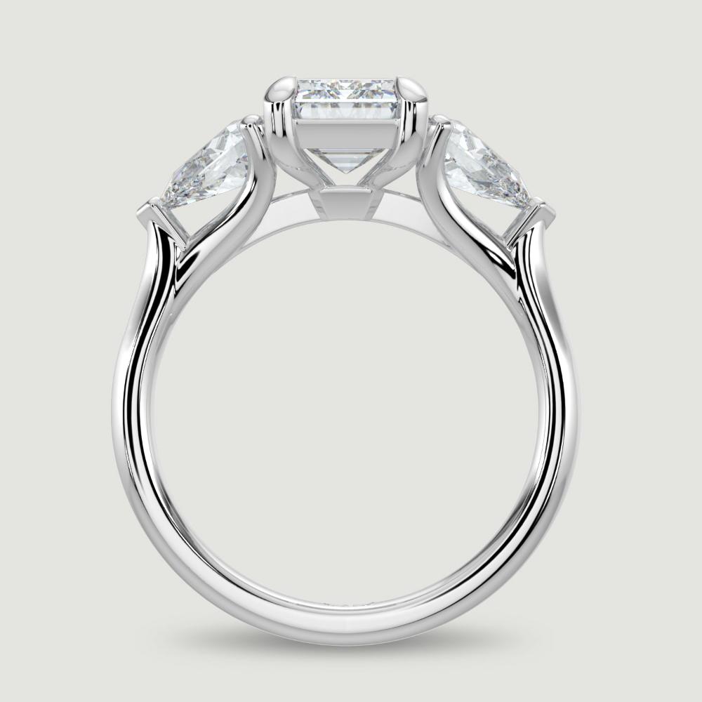 A platinum three stone diamond engagement ring, set with a lab-grown emerald cut diamond centre stone held in a four claw setting flanked by a pear shaped lab-grown diamond on either side, held in three claws. Viewed from the side.
