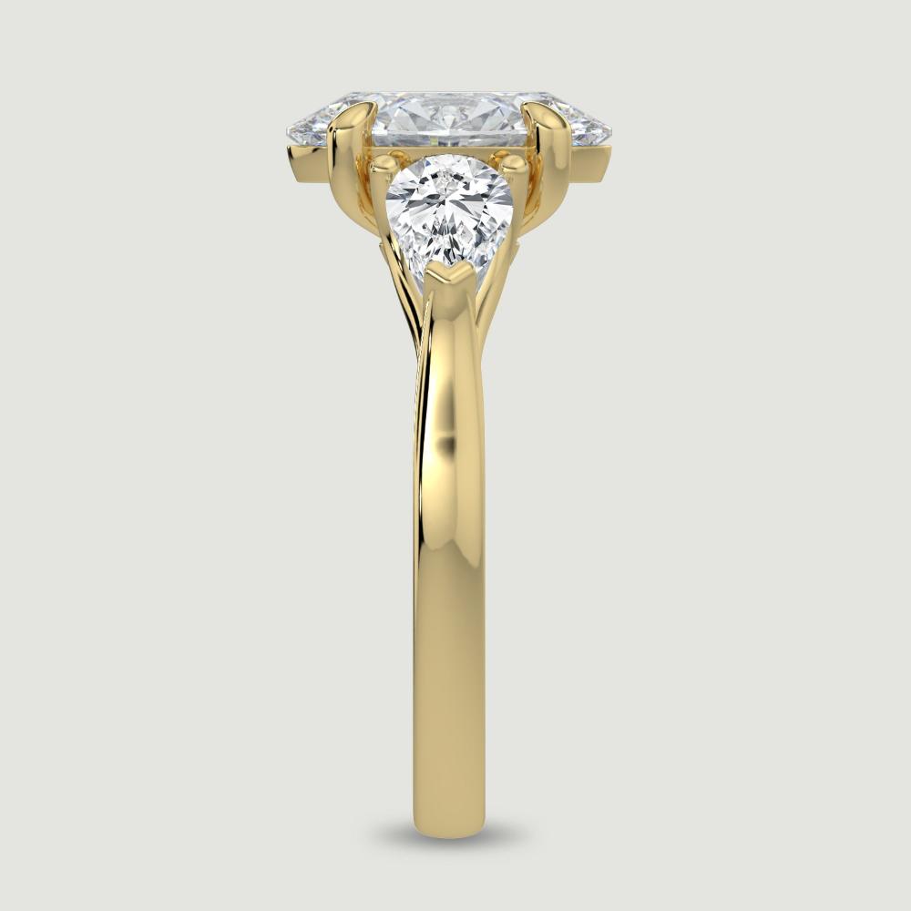 An 18ct yellow gold three stone diamond engagement ring, set with a lab-grown oval shape diamond centre stone held in a four claw setting flanked by a pear shaped lab-grown diamond on either side, held in three claws. Viewed from the edge.