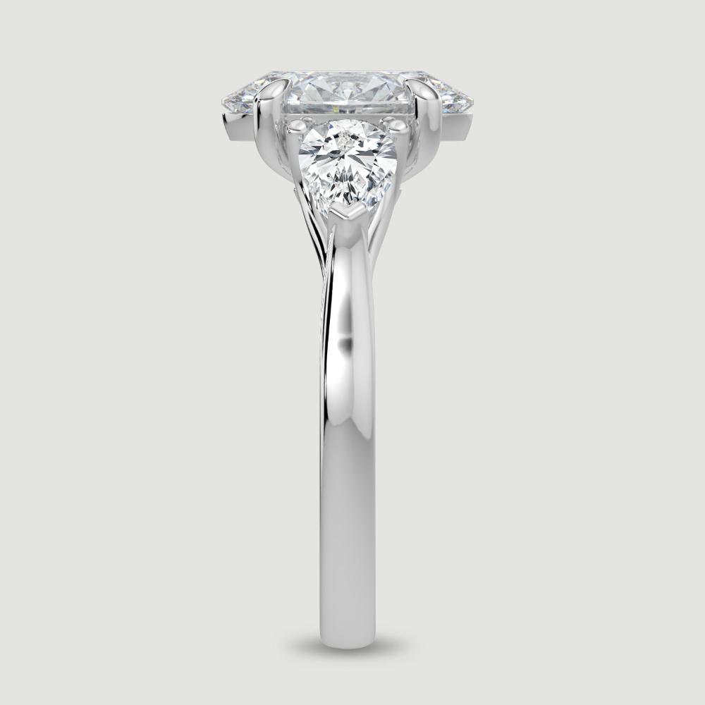 A platinum three stone diamond engagement ring, set with a lab-grown oval shape diamond centre stone held in a four claw setting flanked by a pear shaped lab-grown diamond on either side, held in three claws. Viewed from the edge.