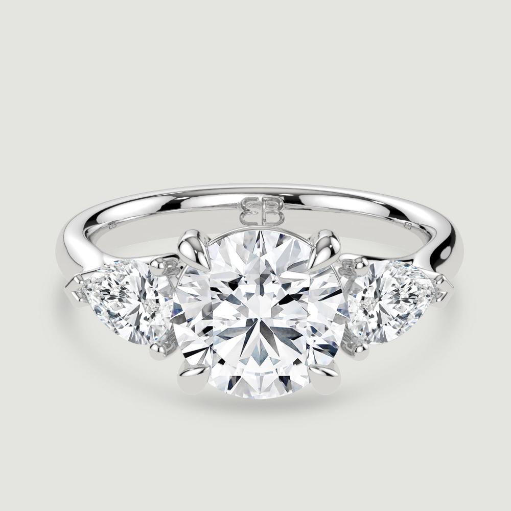 A platinum three stone diamond engagement ring, set with a lab-grown round brilliant diamond centre stone held in a four claw setting flanked by a pear shaped lab-grown diamond on either side, held in three claws. Viewed from above.