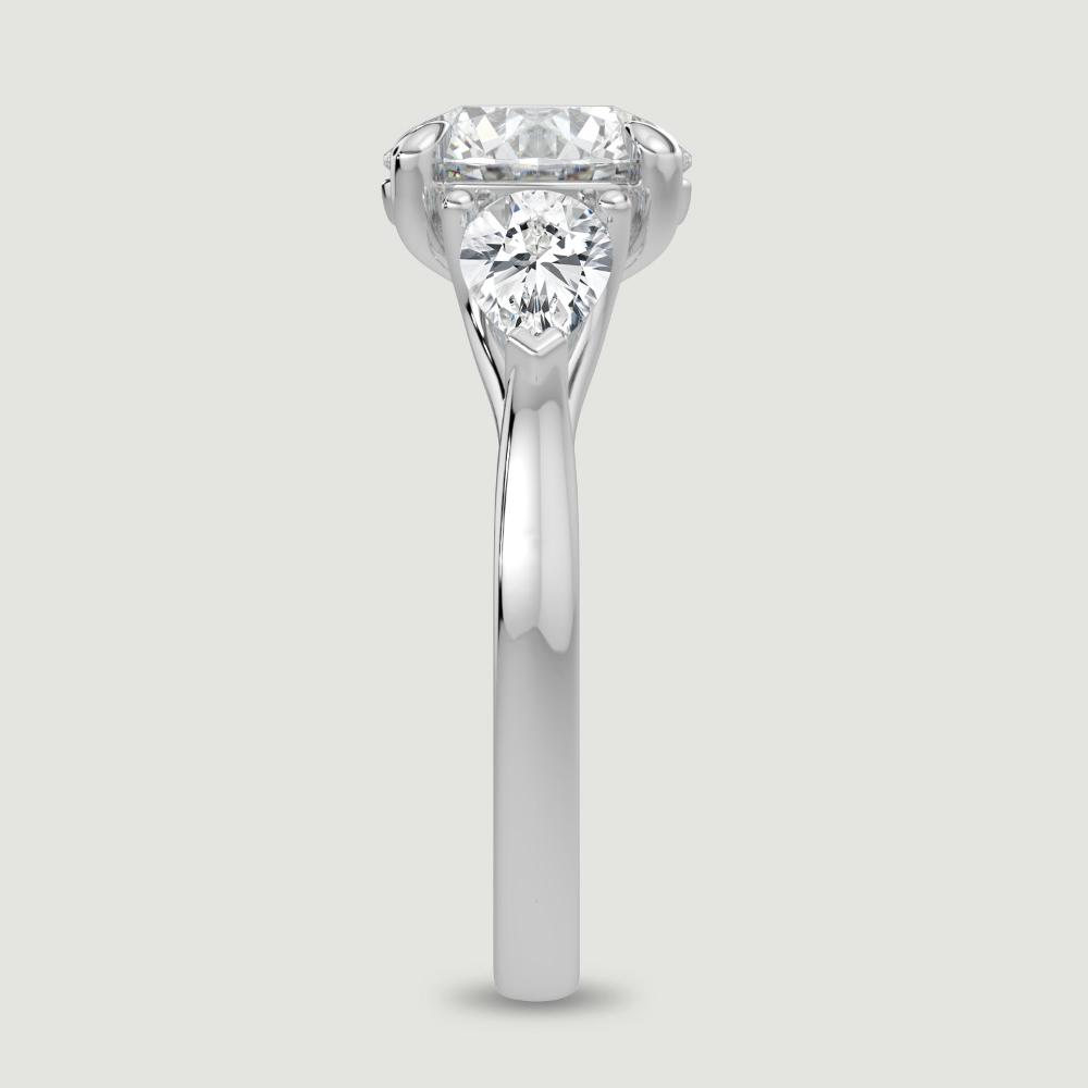 A platinum three stone diamond engagement ring, set with a lab-grown round brilliant diamond centre stone held in a four claw setting flanked by a pear shaped lab-grown diamond on either side, held in three claws. Viewed from the edge.