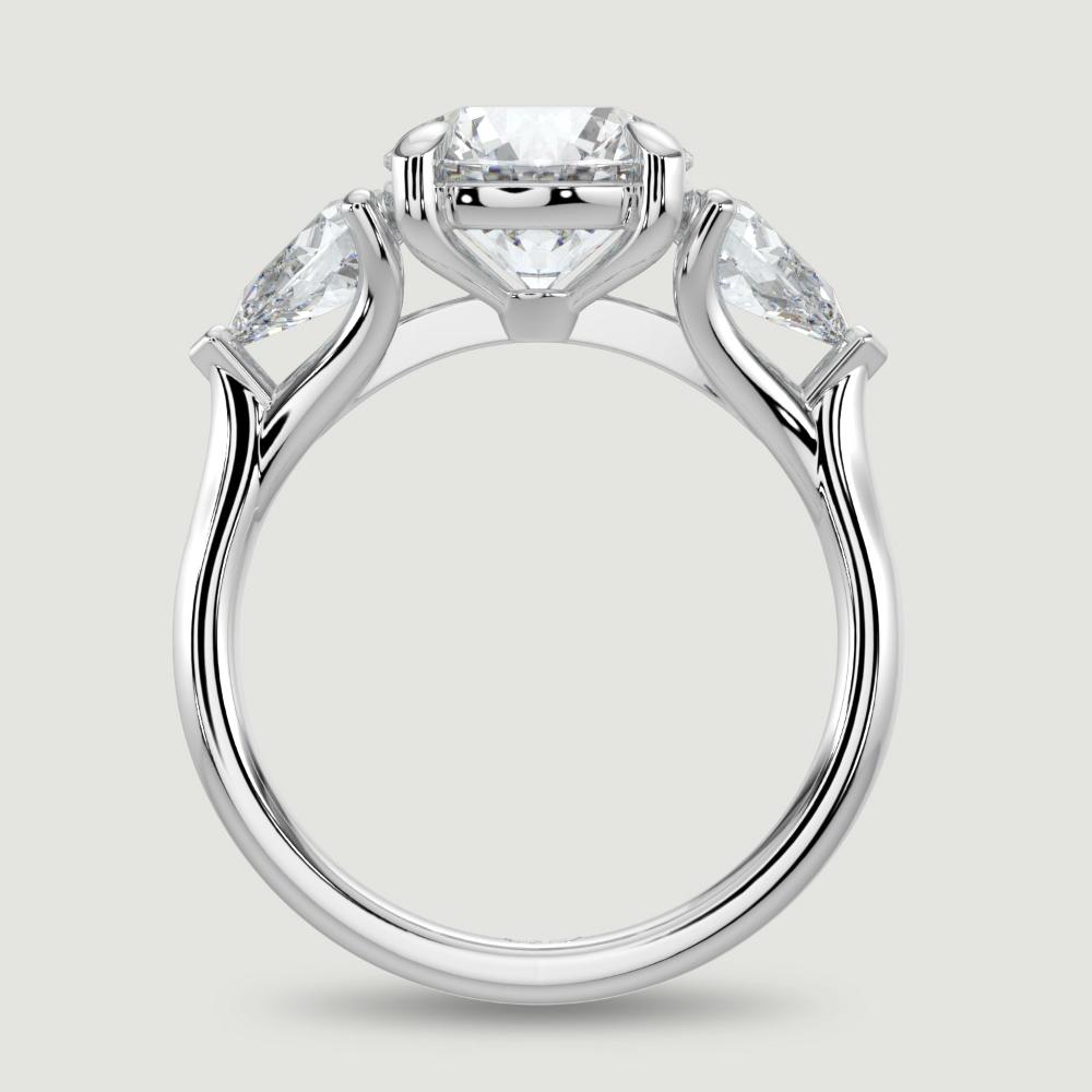A platinum three stone diamond engagement ring, set with a lab-grown round brilliant diamond centre stone held in a four claw setting flanked by a pear shaped lab-grown diamond on either side, held in three claws. Viewed from the side.