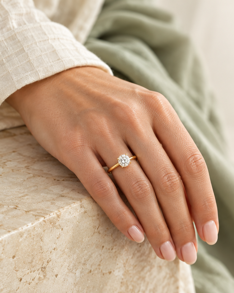 A close-up of a person's hand resting on a stone ledge, wearing a delicate gold ring with a round clear gemstone. The person is dressed in a cream-colored textured top and a sage green blanket, set against a bright, softly lit background.