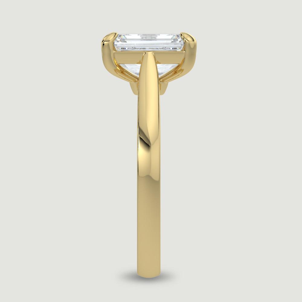 An 18ct yellow gold single stone diamond engagement ring, set with a lab-grown emerald cut diamond held in a four claw setting. Viewed from the edge.
