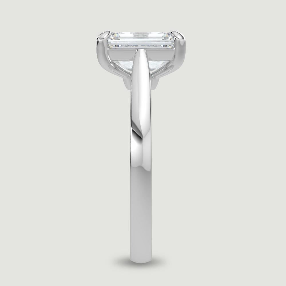 A platinum single stone diamond engagement ring, set with a lab-grown emerald cut diamond held in a four claw setting. Viewed from the edge.