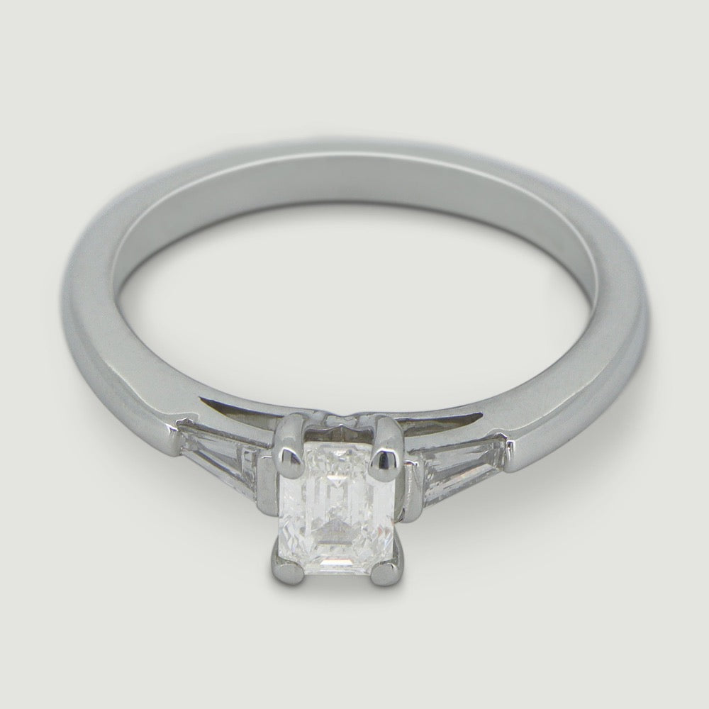A platinum three stone diamond engagement ring. At its heart is a single prominent emerald cut diamond, set in four prongs running down the finger. Set into either side of the rounded band is a tapering baguette cut diamond on either side. Viewed from an angle.