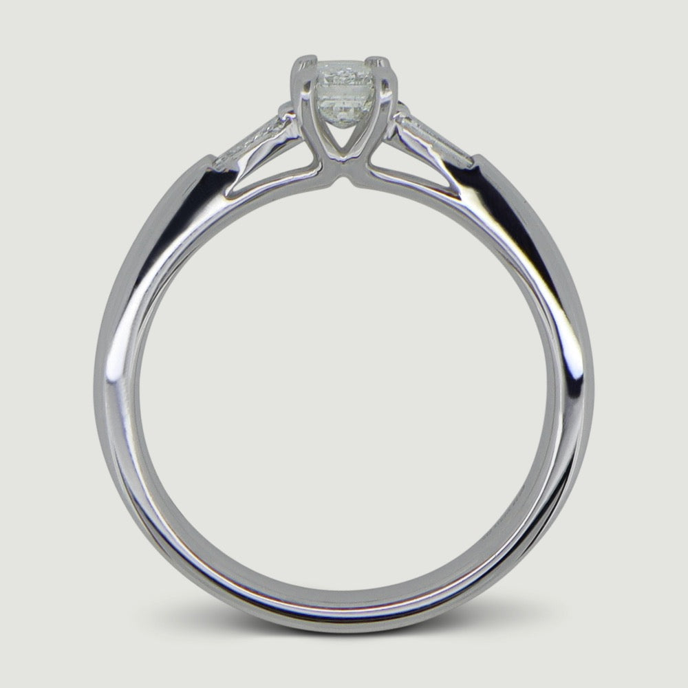 A platinum three stone diamond engagement ring. At its heart is a single prominent emerald cut diamond, set in four prongs running down the finger. Set into either side of the rounded band is a tapering baguette cut diamond on either side. Viewed from the side.