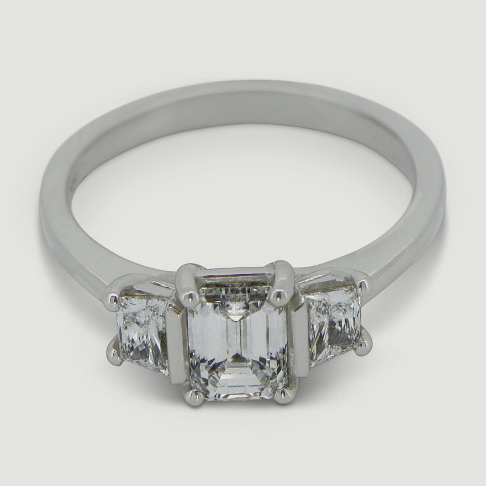 Platinum trilogy diamond ring with an emerald cut centre stone with a trapez cut diamond set on either side - view from an angle
