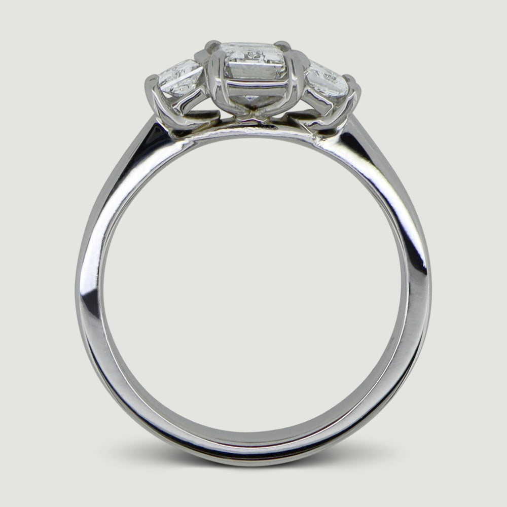 Platinum trilogy diamond ring with an emerald cut centre stone with a trapez cut diamond set on either side - view from the side