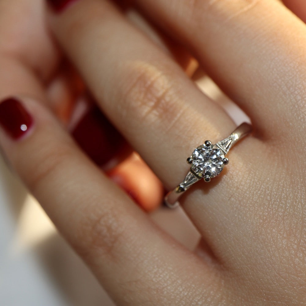 A platinum three stone diamond engagement ring. At its heart is a single prominent round brilliant diamond, set in four prongs running down the finger. Set into either side of the rounded band is a tapering baguette cut diamond on either side. Viewed on the hand.
