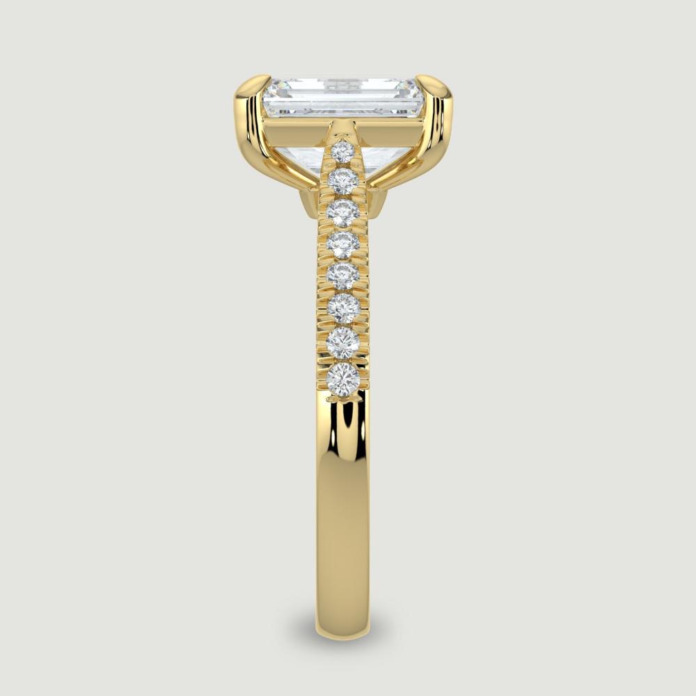 An 18ct yellow gold single stone diamond engagement ring, with micro-pavé set shoulders set with a lab-grown emerald cut diamond held in a four claw setting. Viewed from the edge.