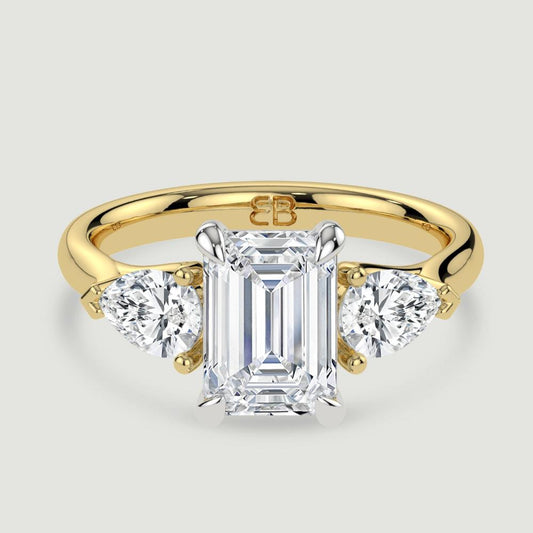 An 18ct yellow gold three stone diamond engagement ring, set with a lab-grown emerald cut diamond centre stone held in a platinum four claw setting flanked by a pear shaped lab-grown diamond on either side, held in three claws. Viewed from above.