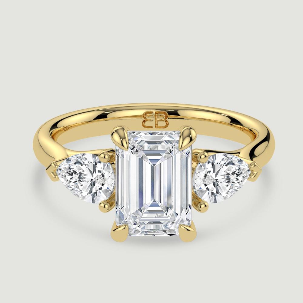 An 18ct yellow gold three stone diamond engagement ring, set with a lab-grown emerald cut diamond centre stone held in a four claw setting flanked by a pear shaped lab-grown diamond on either side, held in three claws. Viewed from above.