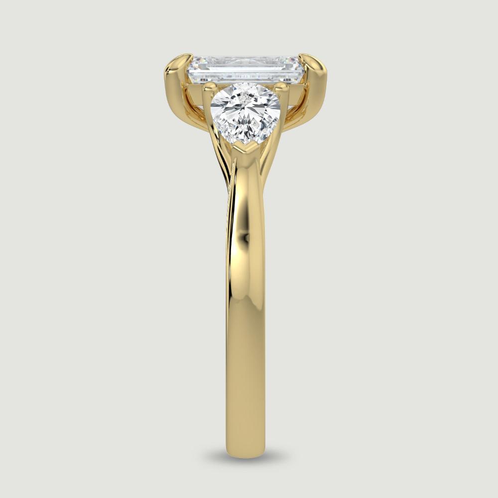 An 18ct yellow gold three stone diamond engagement ring, set with a lab-grown emerald cut diamond centre stone held in a four claw setting flanked by a pear shaped lab-grown diamond on either side, held in three claws. Viewed from the edge.