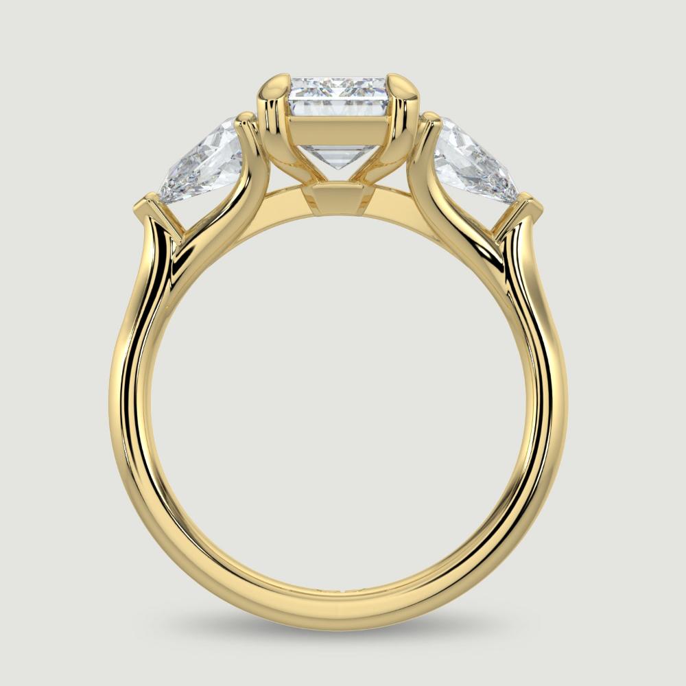 An 18ct yellow gold three stone diamond engagement ring, set with a lab-grown emerald cut diamond centre stone held in a four claw setting flanked by a pear shaped lab-grown diamond on either side, held in three claws. Viewed from the side.