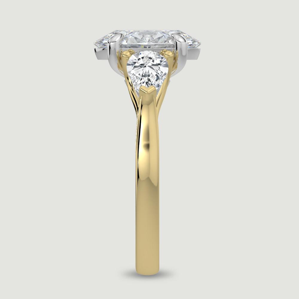 An 18ct yellow gold three stone diamond engagement ring, set with a lab-grown oval shape diamond centre stone held in a platinum four claw setting flanked by a pear shaped lab-grown diamond on either side, held in three claws. Viewed from the edge.