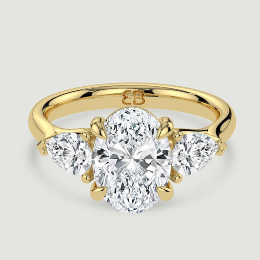 An 18ct yellow gold three stone diamond engagement ring, set with a lab-grown oval shape diamond centre stone held in a four claw setting flanked by a pear shaped lab-grown diamond on either side, held in three claws. Viewed from above.