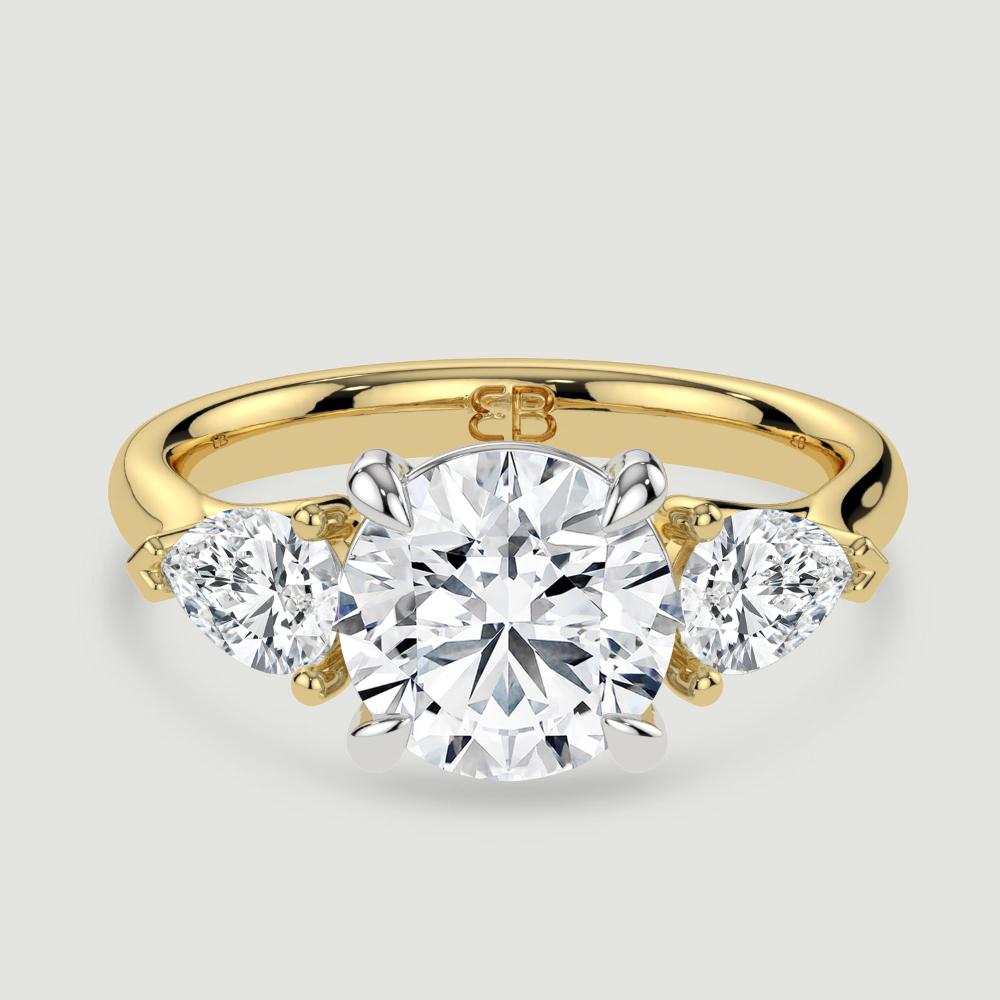 An 18ct yellow gold three stone diamond engagement ring, set with a lab-grown round brilliant diamond centre stone held in a platinum four claw setting flanked by a pear shaped lab-grown diamond on either side, held in three claws. Viewed from above.