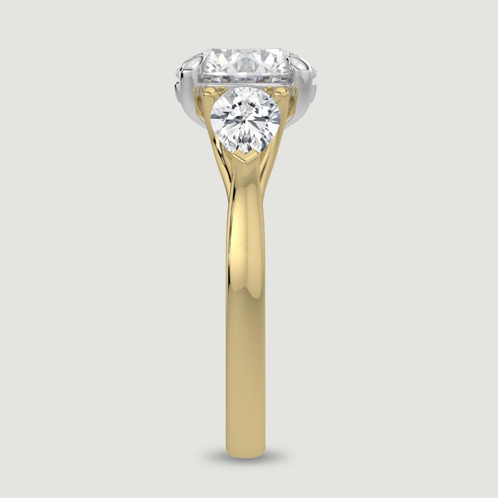 An 18ct yellow gold three stone diamond engagement ring, set with a lab-grown round brilliant diamond centre stone held in a platinum four claw setting flanked by a pear shaped lab-grown diamond on either side, held in three claws. Viewed from the edge.