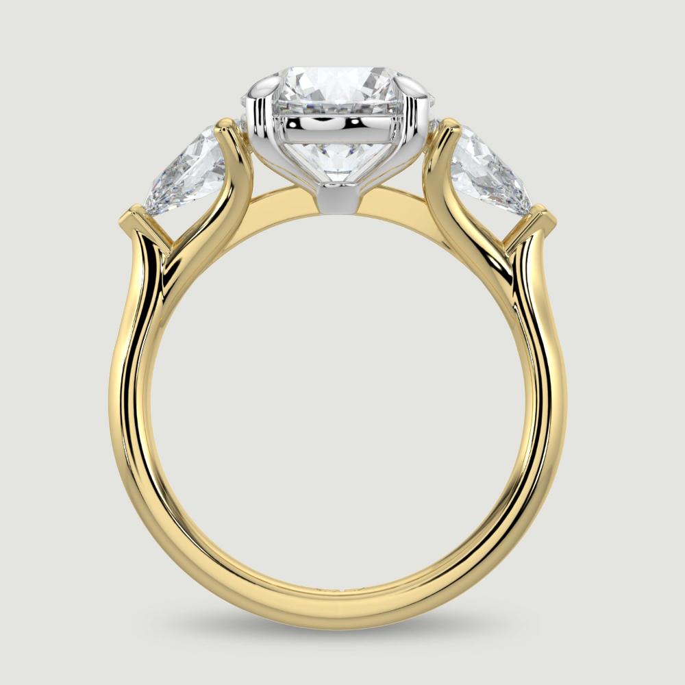 An 18ct yellow gold three stone diamond engagement ring, set with a lab-grown round brilliant diamond centre stone held in a platinum four claw setting flanked by a pear shaped lab-grown diamond on either side, held in three claws. Viewed from the side.