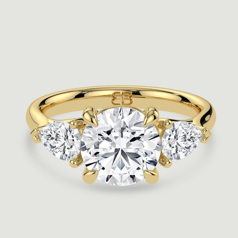 An 18ct yellow gold three stone diamond engagement ring, set with a lab-grown round brilliant diamond centre stone held in a four claw setting flanked by a pear shaped lab-grown diamond on either side, held in three claws. Viewed from above.