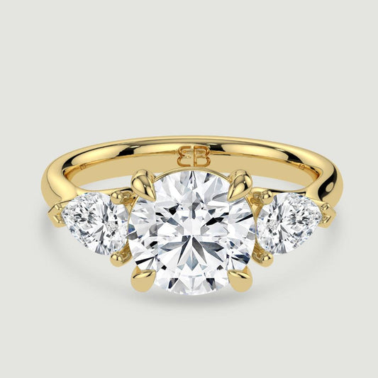 An 18ct yellow gold three stone diamond engagement ring, set with a lab-grown round brilliant diamond centre stone held in a four claw setting flanked by a pear shaped lab-grown diamond on either side, held in three claws. Viewed from above.