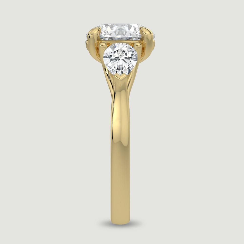 An 18ct yellow gold three stone diamond engagement ring, set with a lab-grown round brilliant diamond centre stone held in a four claw setting flanked by a pear shaped lab-grown diamond on either side, held in three claws. Viewed from the edge.