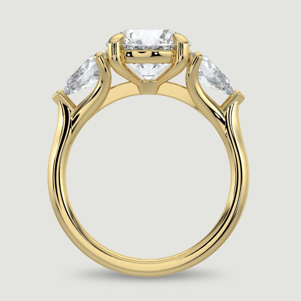 An 18ct yellow gold three stone diamond engagement ring, set with a lab-grown round brilliant diamond centre stone held in a four claw setting flanked by a pear shaped lab-grown diamond on either side, held in three claws. Viewed from the side.