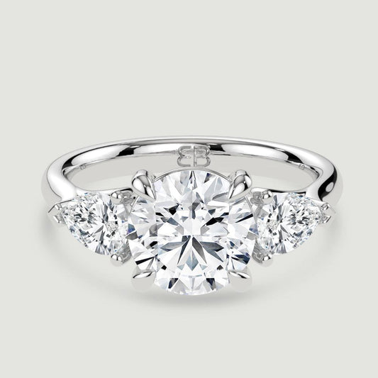 A platinum three stone diamond engagement ring, set with a lab-grown round brilliant diamond centre stone held in a four claw setting flanked by a pear shaped lab-grown diamond on either side, held in three claws. Viewed from above.