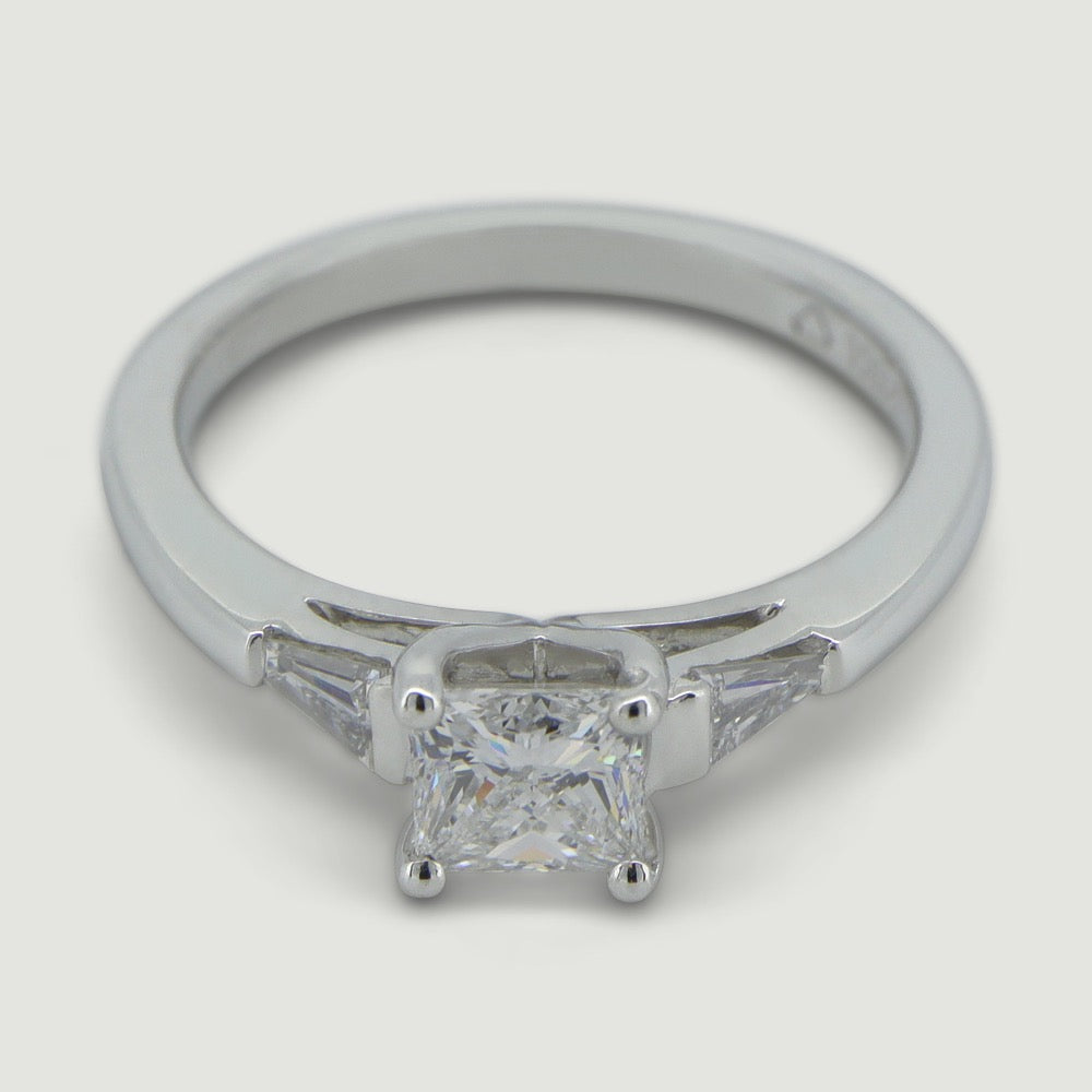 A platinum three stone diamond engagement ring. At its heart is a single prominent princess cut diamond, set in four prongs running down the finger. Set into either side of the rounded band is a tapering baguette cut diamond on either side. Viewed from an angle.