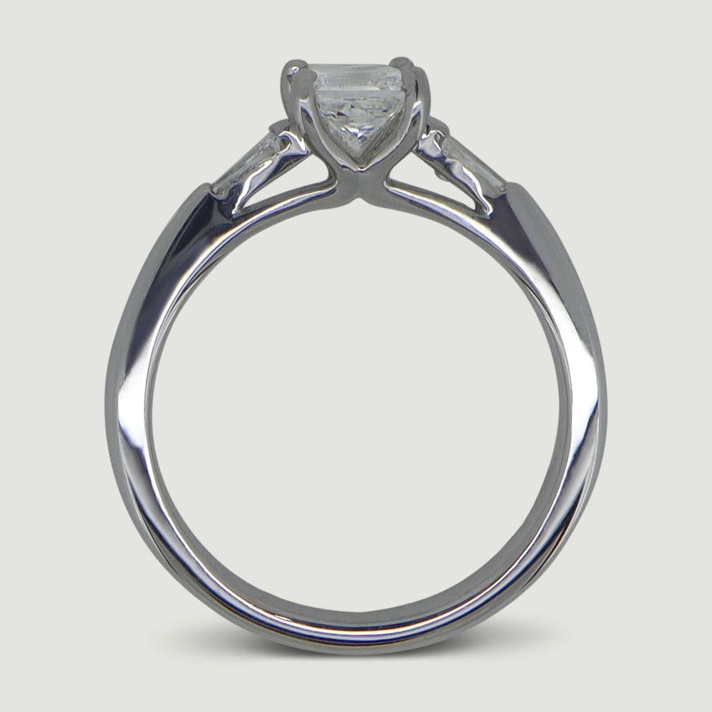 A platinum three stone diamond engagement ring. At its heart is a single prominent princess cut diamond, set in four prongs running down the finger. Set into either side of the rounded band is a tapering baguette cut diamond on either side. Viewed from the side.
