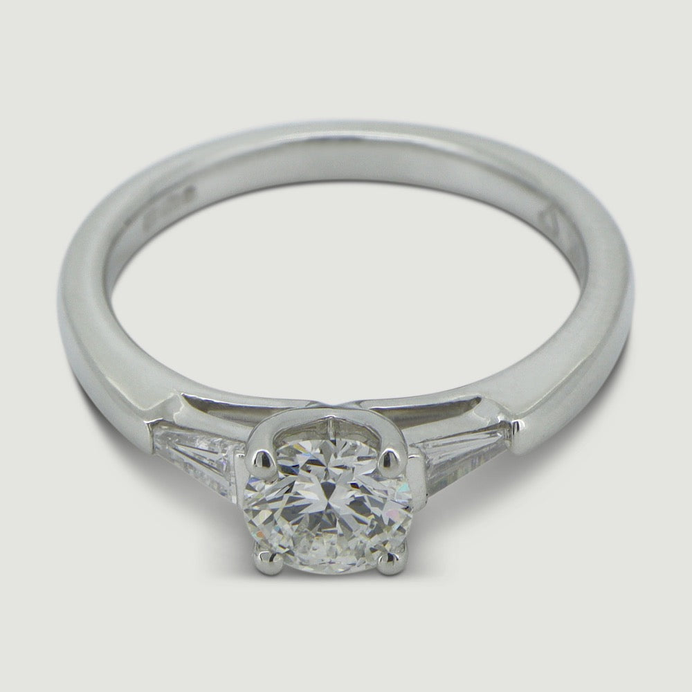 A platinum three stone diamond engagement ring. At its heart is a single prominent round brilliant diamond, set in four prongs running down the finger. Set into either side of the rounded band is a tapering baguette cut diamond on either side. Viewed from and angle.