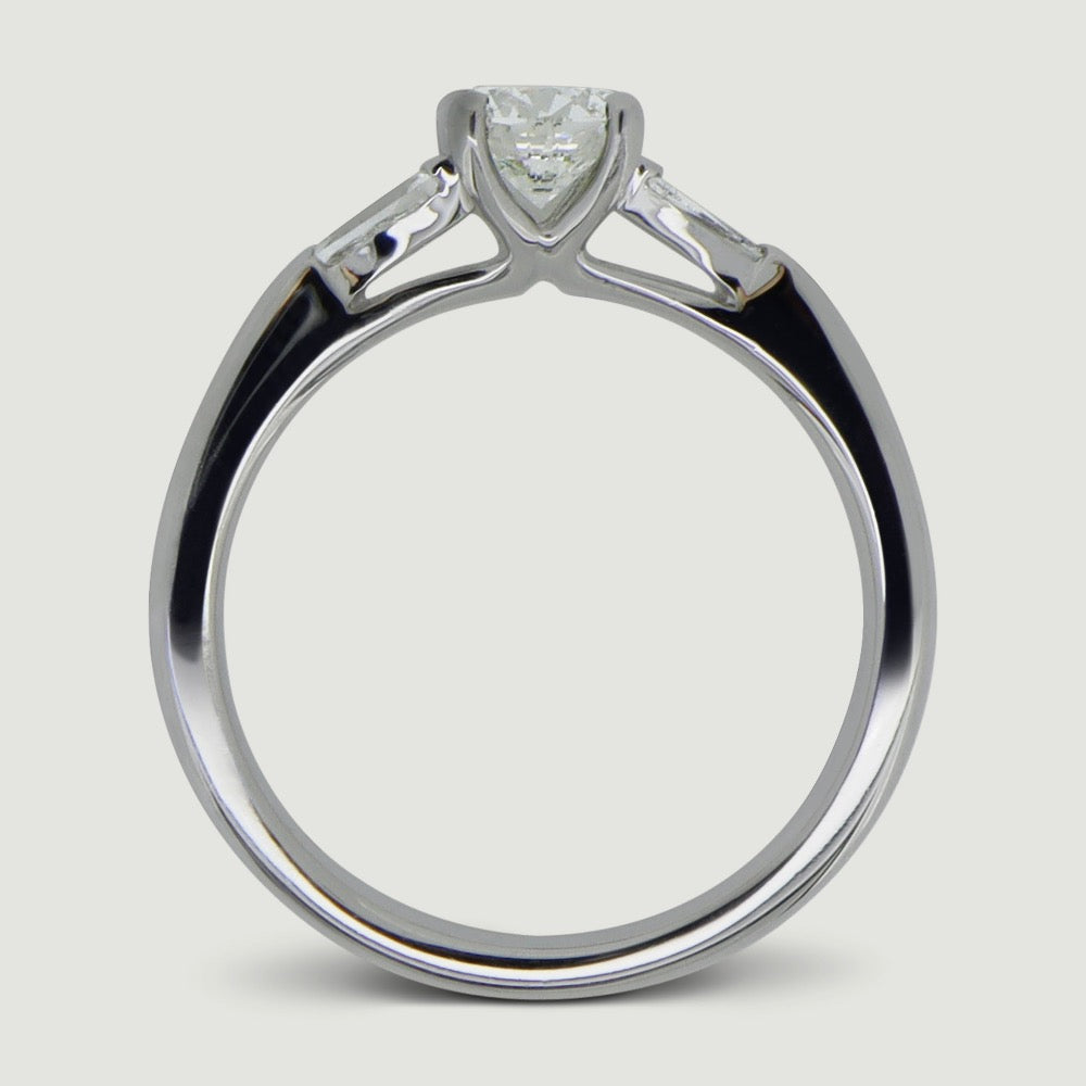 A platinum three stone diamond engagement ring. At its heart is a single prominent round brilliant diamond, set in four prongs running down the finger. Set into either side of the rounded band is a tapering baguette cut diamond on either side. Viewed from the side.